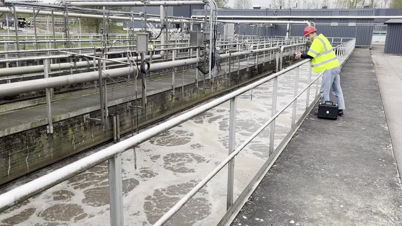 A technician in high-visibility clothing at a wastewater treatment plant, with a carrying case beside the sampling rail
